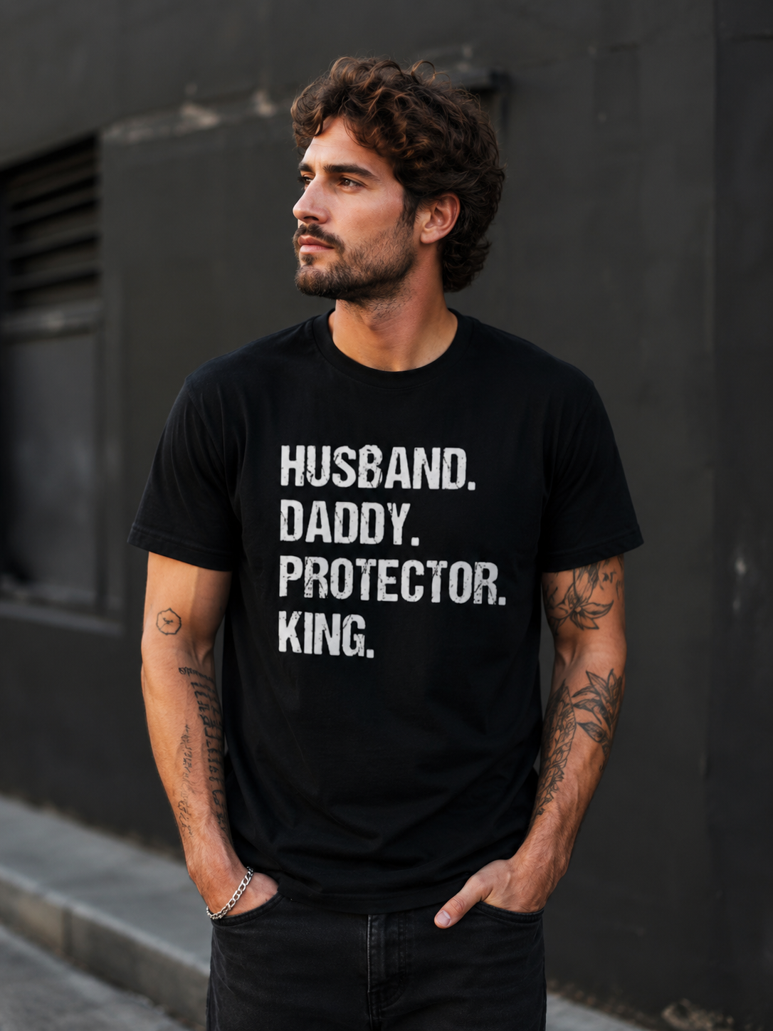 Male model wearing black Husband Daddy Protector King graphic t-shirt featuring distressed white typography in an urban lifestyle setting.